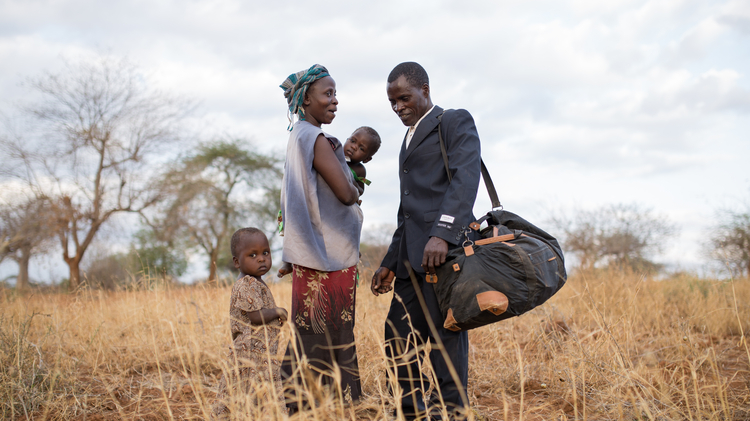 A man in a suit stands with a woman holding a child, while a young child looks on in a dry, grassy field.