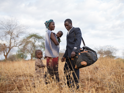 A man in a suit stands with a woman holding a child, while a young child looks on in a dry, grassy field.