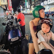 A night scene in a busy street filled with parked scooters, colourful storefronts, and two people on a motorcycle wearing helmets.