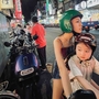 A night scene in a busy street filled with parked scooters, colourful storefronts, and two people on a motorcycle wearing helmets.