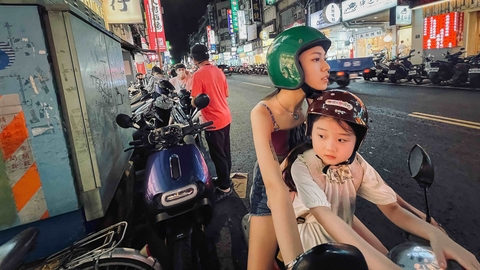 A night scene in a busy street filled with parked scooters, colourful storefronts, and two people on a motorcycle wearing helmets.