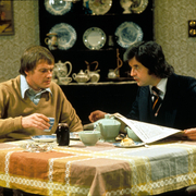 Two men sit at a dining table, sharing tea and food, with decorative china on a shelf in the background and a newspaper on the table.