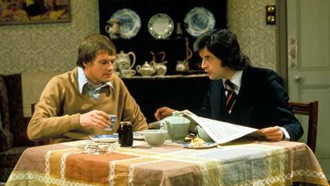 Two men sit at a dining table, sharing tea and food, with decorative china on a shelf in the background and a newspaper on the table.