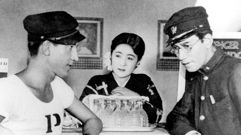 Three individuals interact at a counter displaying glasses; two are in uniforms and one wears a traditional outfit.