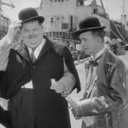 Two individuals in classic vintage attire, including bowler hats and elegant coats, engage near a dock with ships in the background.
