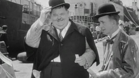 Two individuals in classic vintage attire, including bowler hats and elegant coats, engage near a dock with ships in the background.
