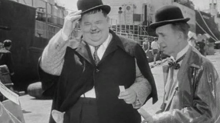 Two individuals in classic vintage attire, including bowler hats and elegant coats, engage near a dock with ships in the background.