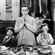 A man and two children are standing with their hands clasped in prayer, set against a dimly lit room.