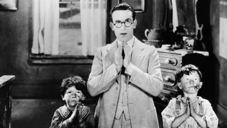 A man and two children are standing with their hands clasped in prayer, set against a dimly lit room.