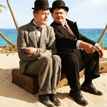 Laurel and Hardy sit on a suitcase on a beach, framed by palm trees and ocean, promoting "Desert Island: The Shorts."
