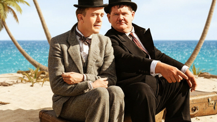 Laurel and Hardy sit on a suitcase on a beach, framed by palm trees and ocean, promoting "Desert Island: The Shorts."