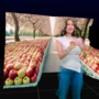 An image of a woman wearing a white t shirt and blue jeans, rendered in a 3D environment, surrounded by two separate shelves of apples, with an apple orchard in the background. She is smiling, looking off camera to the right. 