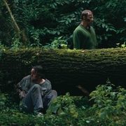 Two individuals stand near a moss-covered fallen tree in a dense forest, surrounded by vibrant greenery and underbrush.