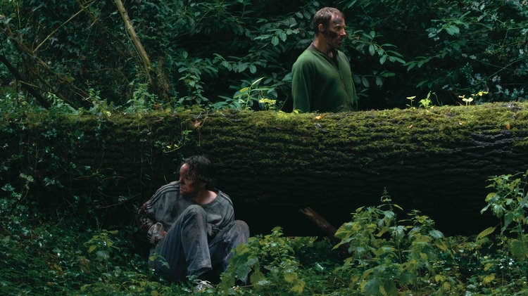 Two individuals stand near a moss-covered fallen tree in a dense forest, surrounded by vibrant greenery and underbrush.