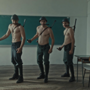 Three shirtless men in military attire stand in a classroom, holding weapons, with a chalkboard that reads "FIUME O MORTE" in the background.