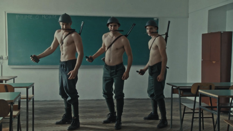 Three shirtless men in military attire stand in a classroom, holding weapons, with a chalkboard that reads "FIUME O MORTE" in the background.