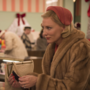 Two women in vintage holiday attire converse in a festive store, surrounded by Christmas decorations and shoppers.