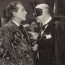 Two men in formal attire engage in conversation, surrounded by classic decor and a bust in the background, exuding a vintage ambiance.