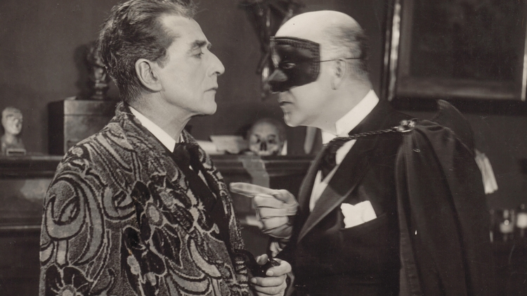 Two men in formal attire engage in conversation, surrounded by classic decor and a bust in the background, exuding a vintage ambiance.