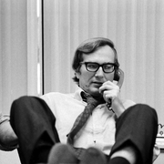 A man casually speaking on the phone with his feet up on a desk, dressed in a white shirt and tie, set in an office with blinds.