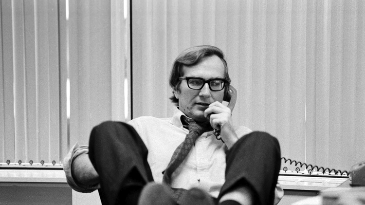 A man casually speaking on the phone with his feet up on a desk, dressed in a white shirt and tie, set in an office with blinds.