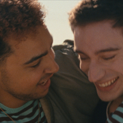 Two friends embrace outdoors, wearing casual clothes with one in a striped shirt and the other in a jacket, amidst a lush, natural setting.