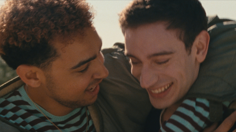 Two friends embrace outdoors, wearing casual clothes with one in a striped shirt and the other in a jacket, amidst a lush, natural setting.