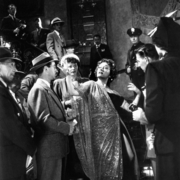 A black-and-white photo depicting a glamorous event, with elegantly dressed people interacting on a grand staircase.