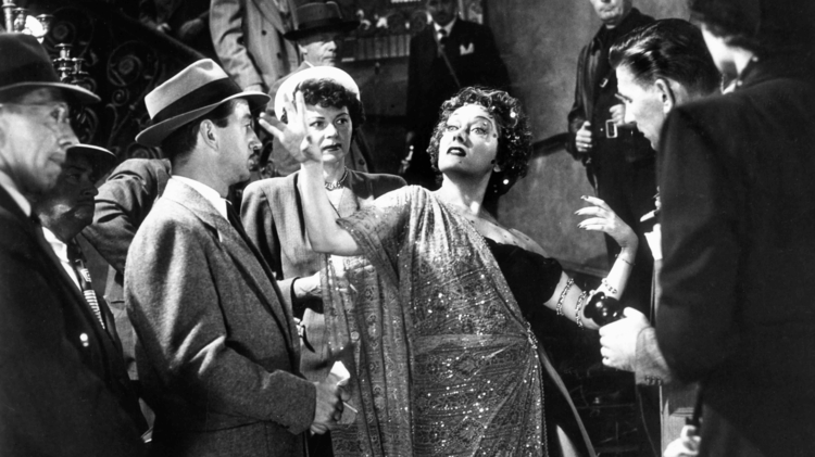 A black-and-white photo depicting a glamorous event, with elegantly dressed people interacting on a grand staircase.