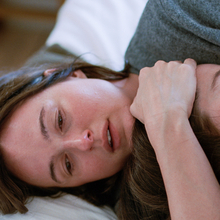 Two people lie on a bed, intertwined in a relaxed manner, showcasing a moment of intimacy and connection.
