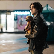 A person in a leather jacket holds a small dog close, standing in an empty nighttime urban setting with glowing shopfronts.