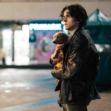 A person in a leather jacket holds a small dog close, standing in an empty nighttime urban setting with glowing shopfronts.