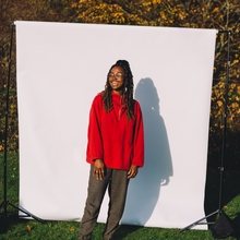 An image of a Emmanuella Blake-Morsi, against a white backdrop situated in an outdoor green space. Emmanuella is a black woman, wearing glasses, a red sweater and dark green trousers, looking off camera to the left, with a smile on her face. 