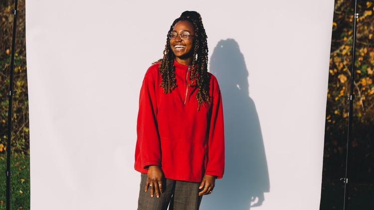 An image of a Emmanuella Blake-Morsi, against a white backdrop situated in an outdoor green space. Emmanuella is a black woman, wearing glasses, a red sweater and dark green trousers, looking off camera to the left, with a smile on her face. 