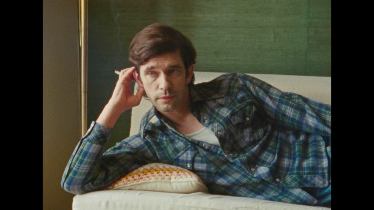 A person in a plaid shirt lounges on a couch, resting their head on one hand against a green wall.