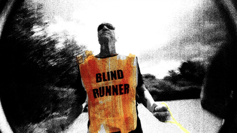 A fisheye camera angle points back at Phil, a blind runner. He wears a bright orange hi-vis tabbard with ‘blind runner’ in large capitals, dark wrap-around sunglasses and clutches a luminous yellow tether that connects him to his guide runner. The whole image is treated with a high-contrast, punk-inspired photocopy texture in black and white, with the accent colours roughly scribbled on top with a highlighter marker.