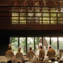 A group of chefs in white uniforms and tall hats work together in a modern kitchen with large windows overlooking greenery.