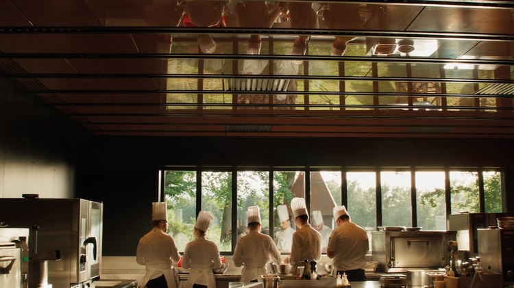 A group of chefs in white uniforms and tall hats work together in a modern kitchen with large windows overlooking greenery.