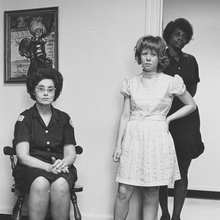 A seated woman in a dark uniform with hands clasped, alongside two girls, one in a white dress, in a room with a painting.