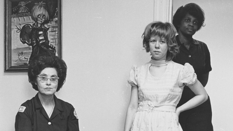 A seated woman in a dark uniform with hands clasped, alongside two girls, one in a white dress, in a room with a painting.