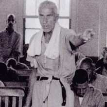 A group of men seated in a room, some listening intently, while one man stands and gestures, appearing to address the others.