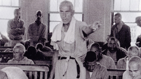 A group of men seated in a room, some listening intently, while one man stands and gestures, appearing to address the others.