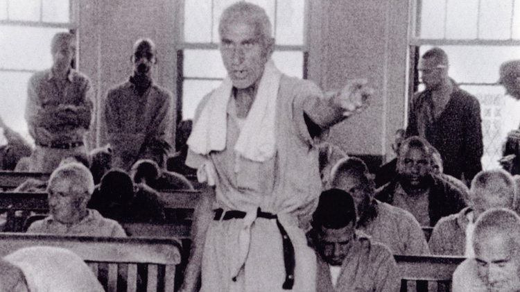 A group of men seated in a room, some listening intently, while one man stands and gestures, appearing to address the others.