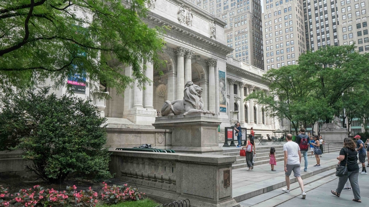 Visitors walk past the New York Public Library, featuring a lion statue and lush greenery amid towering skyscrapers.