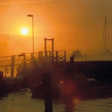 A serene sunset over a misty harbor, silhouetting boats and a dock with a distant sailboat on the water.