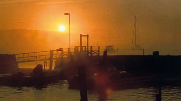 A serene sunset over a misty harbor, silhouetting boats and a dock with a distant sailboat on the water.