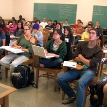 A classroom filled with students seated at desks, some taking notes, others using laptops, engaged in a lecture.