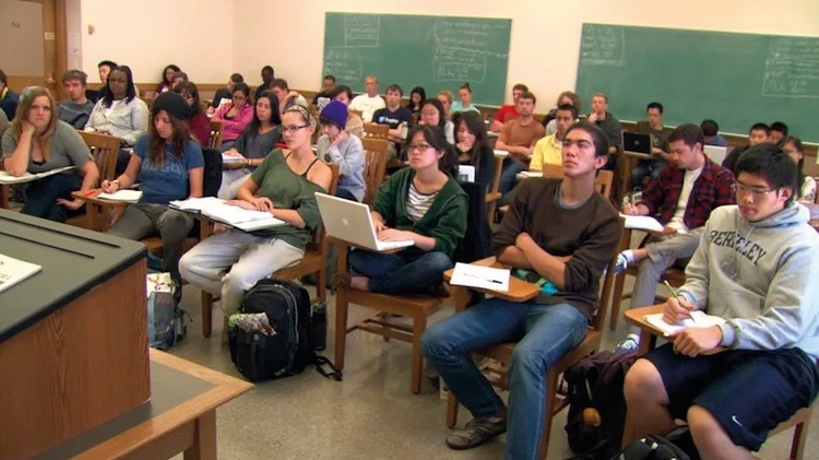 A classroom filled with students seated at desks, some taking notes, others using laptops, engaged in a lecture.