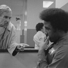 A black-and-white photo depicting two individuals engaged in conversation at a counter in a facility, with others in the background.