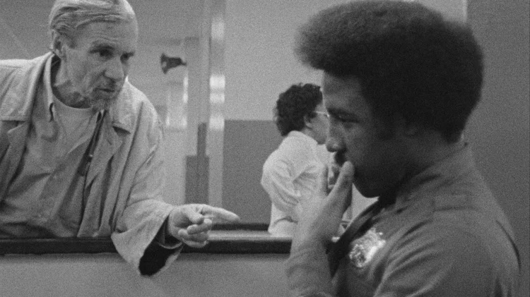 A black-and-white photo depicting two individuals engaged in conversation at a counter in a facility, with others in the background.